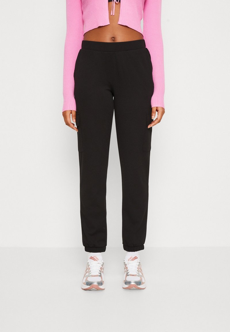Femme portant un pantalon de jogging noir avec poches latérales, un pull court côtelé rose, des chaussettes blanches et des chaussures de sport blanches et roses.