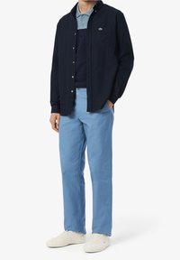 Navy button-up shirt over a blue polo, paired with light blue pants and white sneakers. The shirt features a small green logo on the chest.