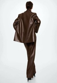 Osoba odchodząca w ciemno-brązowym skórzanym garniturze z szerokimi spodniami i wysokimi obcasami na białym tle.