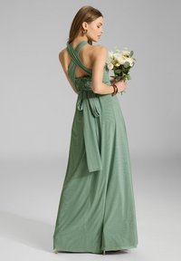 Robe maxi en tissu vert doux, avec un design croisé dans le dos, des accents en dentelle à la taille, une ceinture nouée, et une jupe fluide. Le mannequin tient un bouquet.