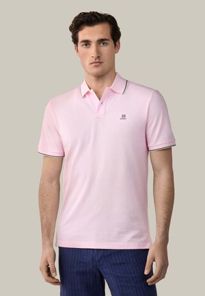 Hombre con camisa tipo polo de manga corta color rosa claro con ribete negro y pantalones azul marino con rayas finas, sobre fondo neutro.