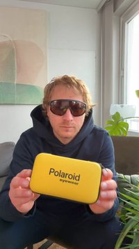 Ryškiai geltona, stačiakampio formos Polaroid akinių dėklas su tekstūruota paviršiaus danga ir juodu logotipu. Juodi akiniai nuo saulės, segimi asmens.