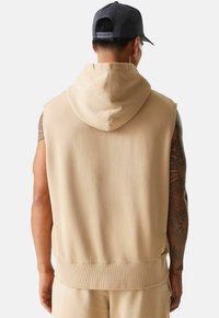 Beige ärmelloses Hoodie mit texturiertem Finish, ausgestattet mit einer Kordelzugkapuze und geripptem Saum. Die Rückansicht zeigt dezente Steppdetails.