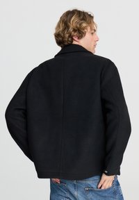 Giacca in misto lana nera con colletto a revers, vestibilità rilassata e polsini con bottone. Presenta un design pulito sul retro senza chiusure visibili.