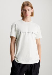 Camiseta blanca de algodón con manga corta y escote redondo. En la parte delantera presenta un texto repetido en negro que dice "Calvin Klein Jeans" y el logotipo.