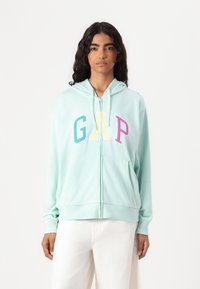 Mintgrøn zip-up hættetrøj lavet af blødt stof, med et stort farverigt "GAP" logo, forlommer og hætte med snøre.