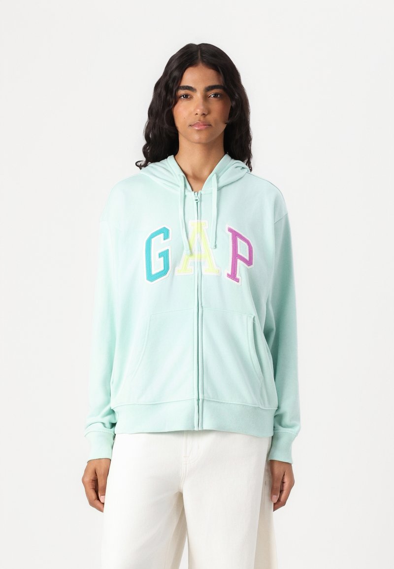 Mintgrøn zip-up hættetrøj lavet af blødt stof, med et stort farverigt "GAP" logo, forlommer og hætte med snøre.