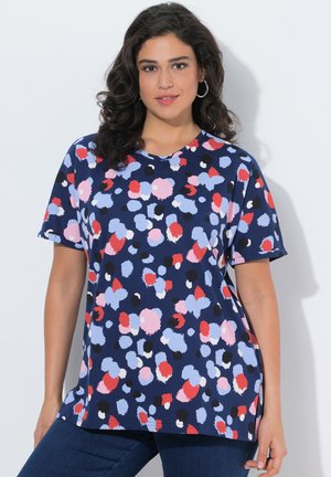 Kobieta o ciemnych kręconych włosach, ubrana w granatową koszulkę z krótkim rękawem ozdobioną abstrakcyjnymi czerwonymi, różowymi, czarnymi i jasnoniebieskimi plamami, pozująca na tle białej ściany.