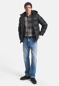 Man som bär svart pufferjacka, grön och röd rutiga skjorta, blå jeans och mörkbruna skor står med händerna i fickorna.