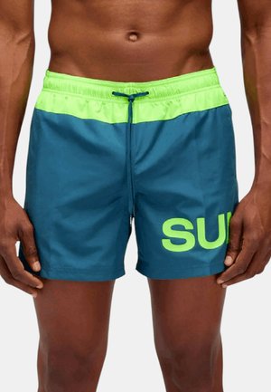 Uomo che indossa pantaloncini da bagno blu con una fascia verde brillante e scritte verdi parziali sulla gamba sinistra.