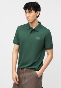 Νεαρός άνδρας που φοράει σκούρο πράσινο πόλο με λογότυπο, καφέ παντελόνι, στέκεται με το ένα χέρι στην τσέπη σε απλό φόντο.