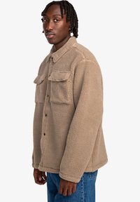 Tan fleece jacket with a folded collar, front buttons, and two chest pockets. The fabric has a fluffy texture. Worn with blue jeans.