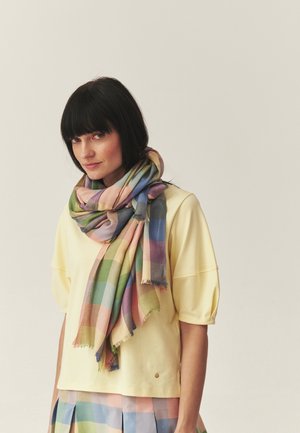 Woman with short black hair wearing a yellow shirt, multicolored plaid scarf, and matching plaid skirt, standing against a plain background.