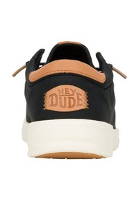 Sneaker nera in tela con accento in pelle color tan sul retro, caratterizzata da un logo circolare "HEY DUDE", suola bianca e design leggero.
