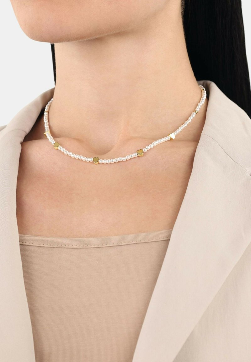 Collana di perle con perle bianche e accenti a forma di cuore dorati, progettata con una texture sottile e liscia, indossata sopra un top beige.