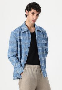 Blauwe geblokte denimjas met onafgewerkte randen, knoopsluiting en een ontspannen pasvorm, gecombineerd met een zwart shirt en beige broek.