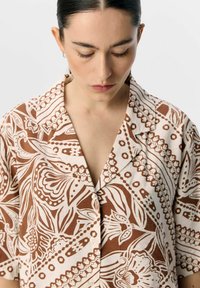Camicia a maniche corte di colore marrone e bianco con un colletto e chiusura a bottoni. Il tessuto ha un aspetto testurizzato e un motivo floreale.