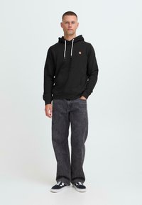 Giovane in piedi, indossa un hoodie nero con lacci bianchi, jeans neri sbiaditi e sneakers nere con suola bianca.