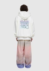 Vit hoodie med färggrafisk text på baksidan, parat med oversized gradientbyxor som övergår från rosa till blått. Mjuk tygtextur.