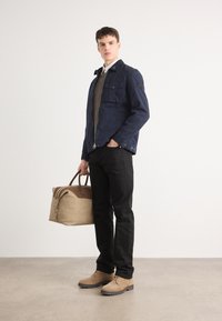 Casaco azul-marinho com vários bolsos, suéter de malha bege, calças pretas, sapatos bege e uma mala bege com detalhes castanhos. Texturas suaves em todo o conjunto.