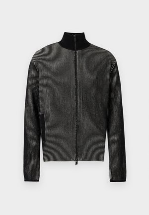 Joven con chaqueta negra de rayas con cremallera, gorra negra, pantalones cortos negros anchos, calcetines negros y deportivas negras gruesas sobre un fondo liso.