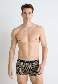 Schwarze und goldene gemusterte Boxershorts mit einem einfarbigen schwarzen Bund, der mit "BOSS" gebrandet ist. Der Stoff wirkt glatt und hat eine figurbetonte Passform.