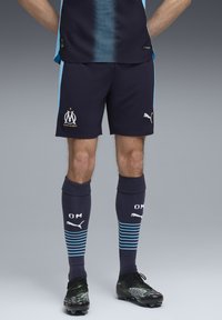 Mørkeblå sports shorts med lyseblå accenter, logo og tekst. Sorte fodboldsokker med hvid tekst og blå striber. Sorte fodboldstøvler med mønster.