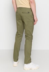 Hombre con pantalones ajustados verde olivo y zapatillas blancas, de pie contra un fondo claro y liso, vista desde atrás.