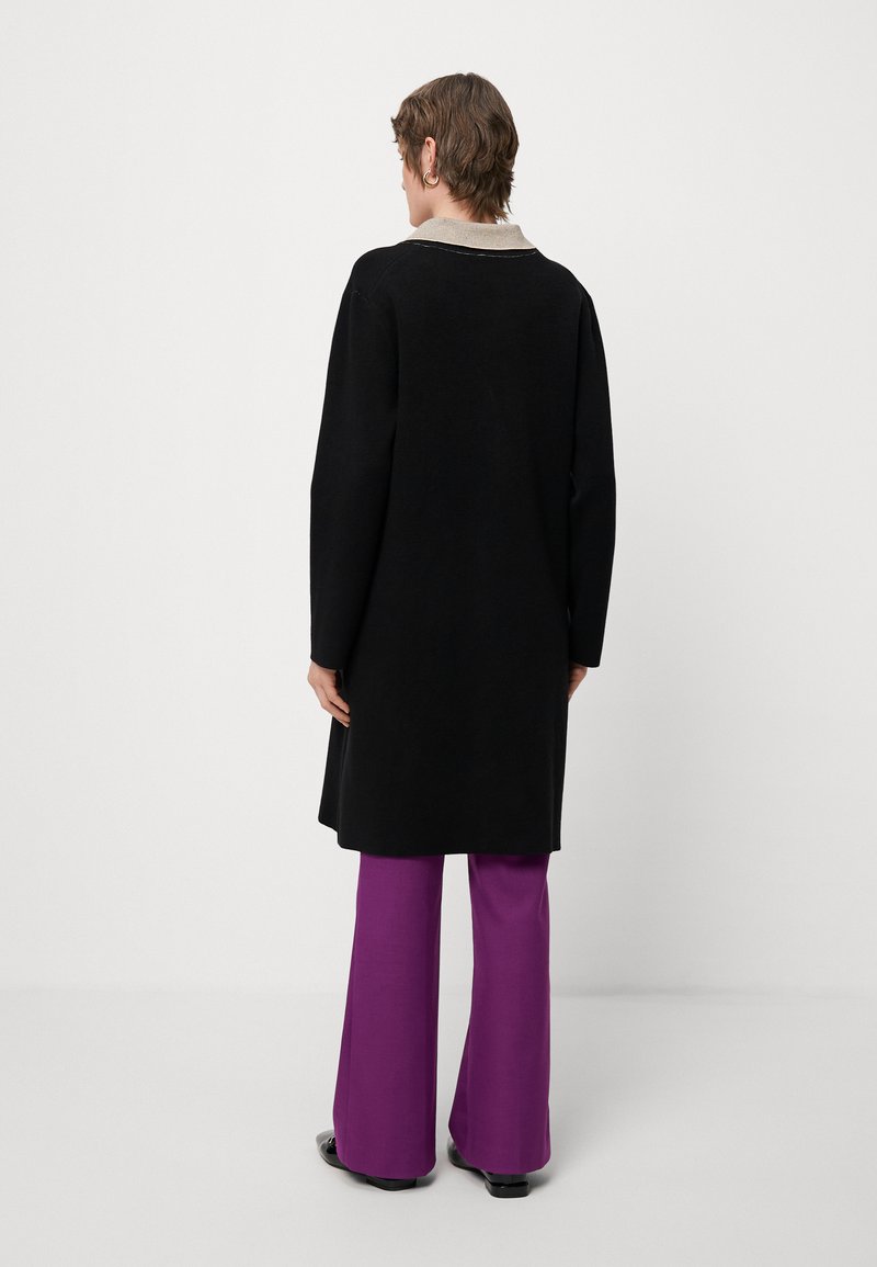 Cappotto lungo nero a maniche lunghe con colletto leggero, abbinato a pantaloni viola svasati. Il cappotto presenta un design semplice e una texture liscia.