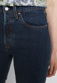 Jeans de mezclilla azul oscuro con un diseño clásico de cinco bolsillos, herrajes de cobre y costuras amarillas contrastantes, que exhiben una textura suave.