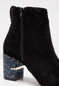 Bottine en suédine noire avec une finition texturée, dotée d'une fermeture éclair sur le côté et d'un talon carré recouvert de paillettes multicolores.