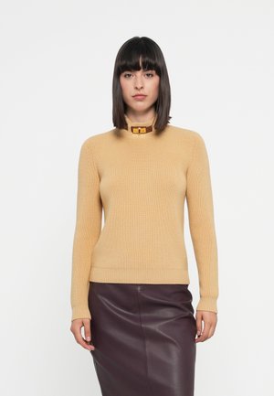 Beige ribbed knit sweater with a high neckline and a gold buckle accent. Features long sleeves and a fitted silhouette. Dark purple leather skirt.