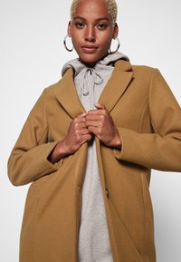 Cappotto cammello realizzato in tessuto morbido, con colletto a revers, chiusura frontale con bottoni e due tasche laterali, indossato sopra un hoodie grigio.