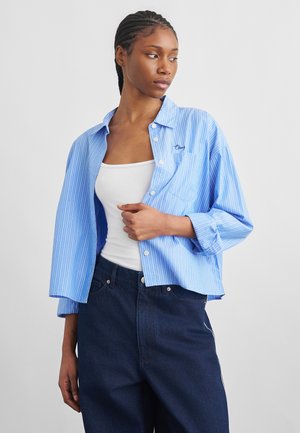 Žena s vrkočmi, oblečená v otvorenej svetlomodrej pruhovanej košeli cez biele tielko a tmavomodré džínsy, pozerá sa doľava.