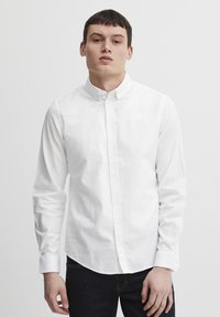 Camisa blanca de botones, con mangas largas, confeccionada en algodón, con cuello clásico y una textura suave, combinada con jeans de mezclilla oscura.