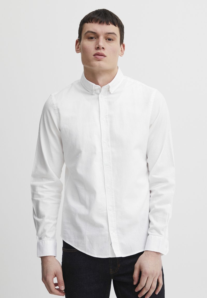 Camisa blanca de botones, con mangas largas, confeccionada en algodón, con cuello clásico y una textura suave, combinada con jeans de mezclilla oscura.