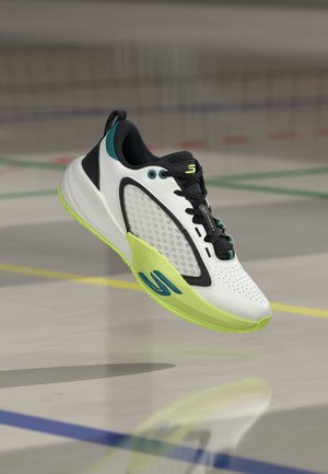 Chaussure de sport blanche avec des détails noirs et jaune fluo flottant au-dessus d’un sol de gym marqué de lignes colorées.