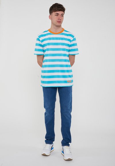 T-shirt a maniche corte a righe verticali in blu e bianco con colletto arancione, abbinata a jeans denim blu e scarpe bianche.
