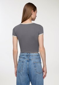 Kobieta z prostymi brązowymi włosami, ubraną w dopasowany szary top z krótkim rękawem wykończony beżowym obszyciem oraz wysoko taliowane niebieskie dżinsy, pokazana od tyłu.