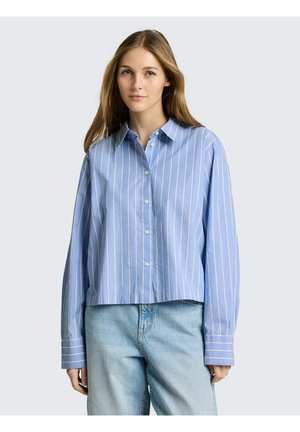 Junge Frau trägt ein locker sitzendes, hellblaues Hemd mit weißen vertikalen Streifen und hellblaue Jeans, steht vor einem schlichten Hintergrund.