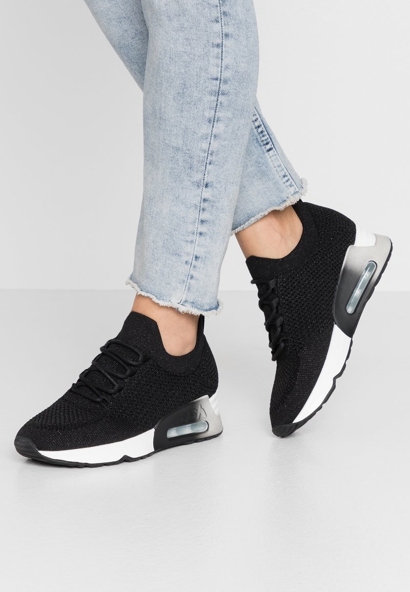 Personne portant des baskets en maille noire avec semelles blanches et talons à coussin d'air, associées à un jean bleu clair à ourlet effiloché, sur un sol blanc.