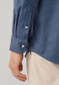 Blauw herringbone overhemd met een list, knoopmanchetten en een gebogen zoom. De stof heeft een lichte textuur, gecombineerd met lichtgekleurde broeken.