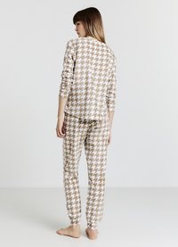 Completo pigiama beige con motivo pied-de-poule; maglia a maniche lunghe e pantaloni aderenti; tessuto morbido e strutturato; piedi nudi visibili, sfondo neutro.