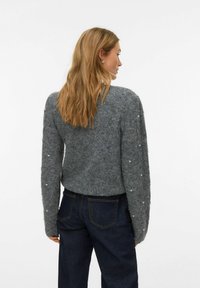 Šedý pletený svetr s dlouhými rukávy, zdobený perličkami na rukávech, měkkou strukturou a volnějším střihem. Kombinován s tmavými džínami.