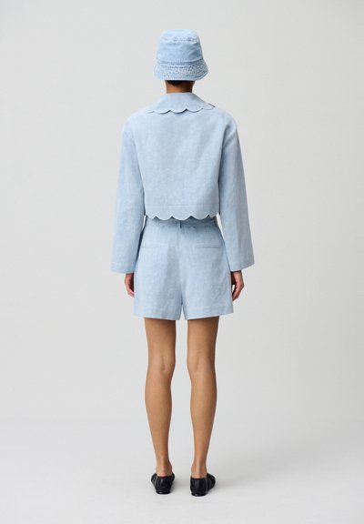 Chaqueta cropped de lino azul claro con cuello con festón, combinada con pantalones cortos a juego. Lleva puesto un sombrero de cubo azul claro y zapatos planos negros.