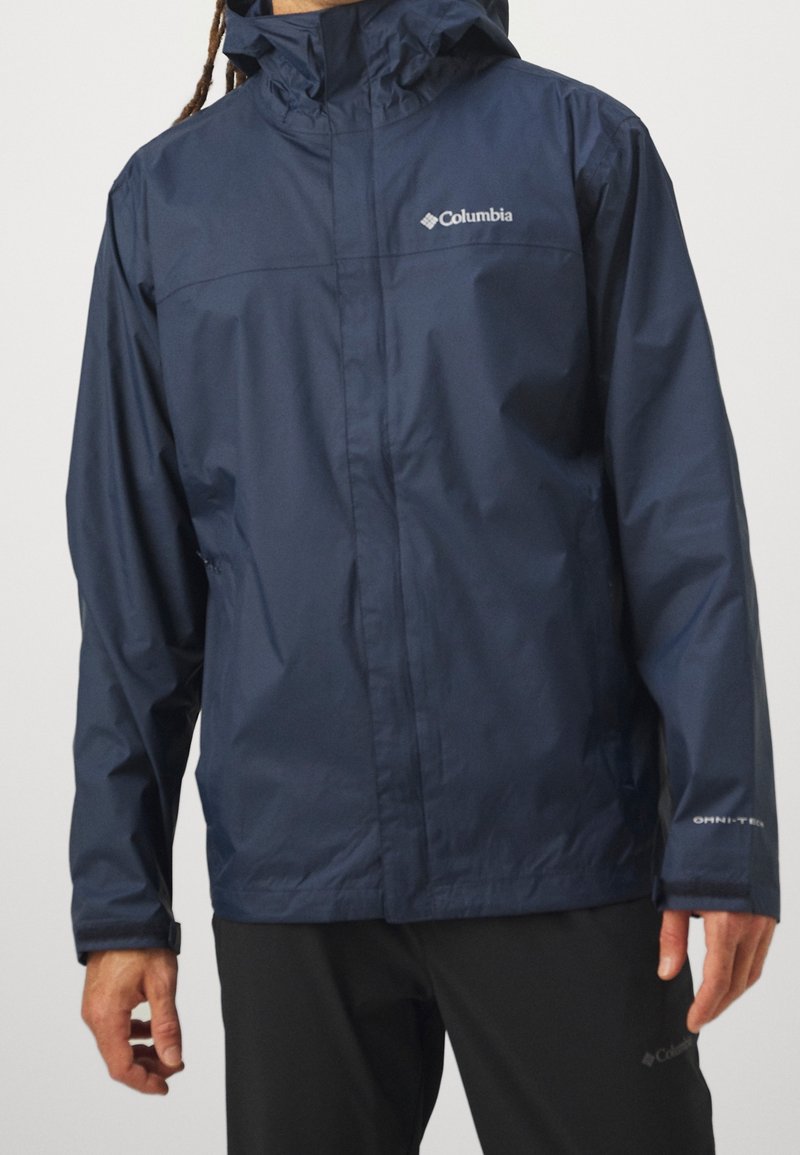 Homme portant une veste imperméable Columbia bleu marine avec capuche et un pantalon noir, debout devant un fond clair uni.