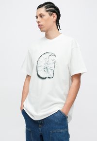 Biele bavlnené tričko s veľkým abstraktným čierno-šedým grafickým dizajnom na prednej strane. Voľný strih, krátke rukávy, okrúhly výstrih. Modré riflové šortky.