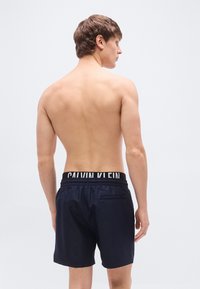 Μπλε σκούρο ανδρικό μαγιό βερμούδα με ελαστικ ή μέση και λευκό λογότυπο Calvin Klein. Διαθέτει πίσω τσέπη και απαλό, ελαφρύ υλικό.