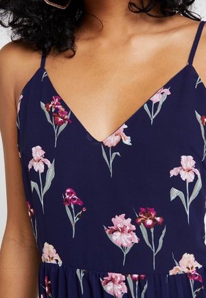 Kvinde iført en mørkeblå kjole med tynde stropper og et blomstermønster med pink og lilla blomster.