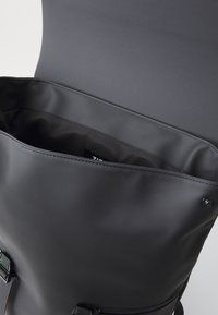 Mochila negra tipo roll-top con acabado mate, que cuenta con un interior amplio, textura suave y correas ajustables. Incluye un cierre a presión.